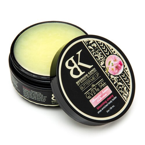 An image of a round container of Butter Me Up in the Blooming Cherry scent. The container is open with the lid propped up in front of it, revealing the smooth pale yellow surface of the body butter.
